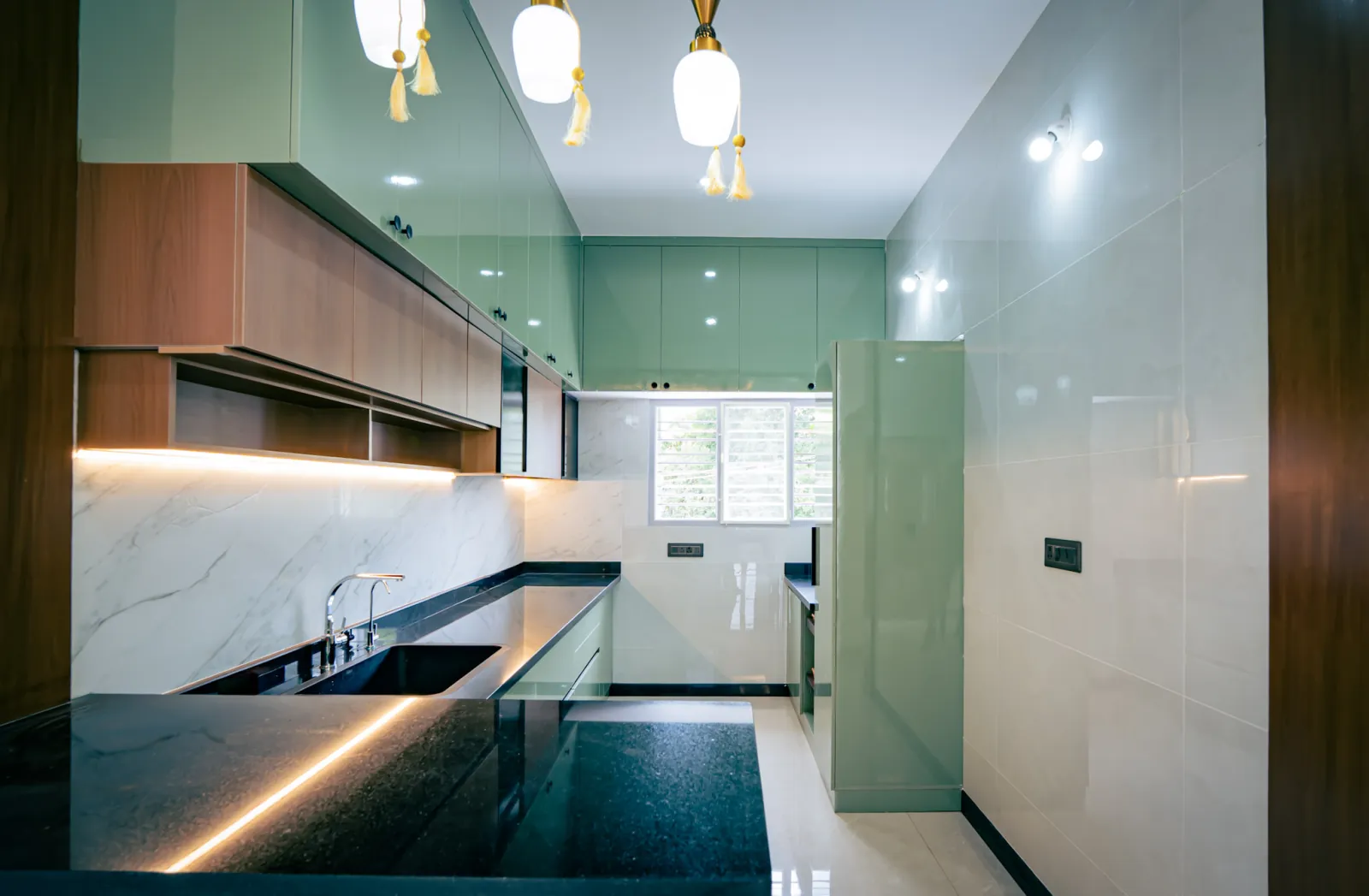 Sage-green modular kitchen with wood-finish wall cabinets, under-cabinet lighting and a black granite counter